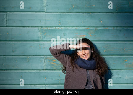 Asiatische Mädchen steht an der Wand und kniff ihre Augen wegen der hellen Sonne. Sie lächelt und versucht, ihr Gesicht mit der Hand abdecken. Stockfoto