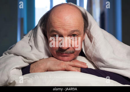 Ein älterer glatzköpfiger Mann mit einem Schnurrbart liegt im Bett, eine Kissen umarmt. Er hat ein Problem mit dem Schlaf. Stockfoto