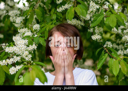 Ein schönes Mädchen steht der blühenden Vogel-Kirsche und niest. Sie leidet an einer Allergie bis zur Blüte im Frühjahr. Stockfoto