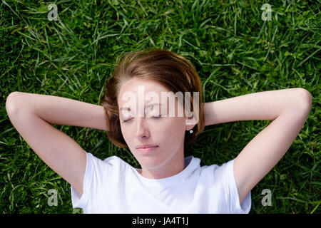 Ein schönes Mädchen mit Sommersprossen liegt auf dem grünen Rasen mit geschlossenen Augen und Pausen Stockfoto