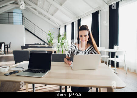 Porträt von einem Designer arbeiten Zuhause auf neue Ideen. Eine dunkles Haar geflochten Frau sitzt an einem weißen Tisch in Freizeitkleidung, sucht sie in die Kamera, mit einem Bleistift, mit einem Laptop vor ihr. Stockfoto