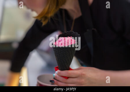 Junge Verkäuferin in einer Eisdiele setzen helle Rosa Frucht Eis Kugeln in schwarz Waffelhörnchen. Tiefenschärfe für Nutzung im Hintergrund. Stockfoto