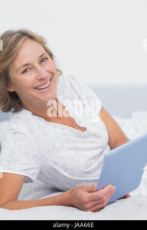 Lächelnde blonde Frau auf Bett mit TabletPC Blick in die Kamera im Schlafzimmer zu Hause Stockfoto
