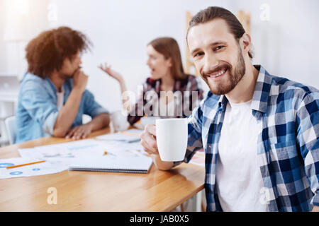 Tolle Leute, tolle Firma. Schön entspannt geselliger Mann am Tisch sitzen und eine Tasse mit Heißgetränk während seiner Kollegen diskutieren etwas halten Stockfoto