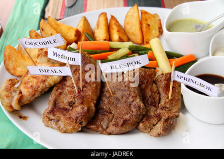 Ein Teller mit afrikanischen Spiel Fleisch mit Saucen, Gemüse und Kartoffel Pommes.  Fleisch sind mit kleinen Zahnstocher Schilder gekennzeichnet; Krokodil, Springbock, Impala, Blessböcke Stockfoto