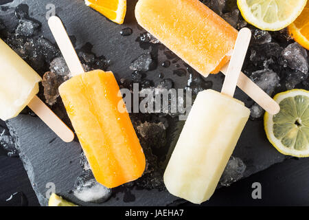 Hausgemachte Orange und Zitrone Eis am Stiel mit Eis und Zitrusfrüchte auf Stein schwarzem Hintergrund. Sommer-Food-Konzept. Ansicht von oben. Stockfoto