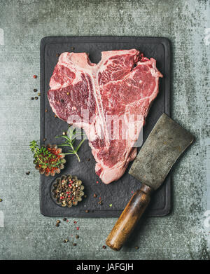 Frisches rohes Rindfleisch roh t-Bone Steak mit Gewürzen auf schwarzem Schiefer Steinplatte über graue konkreten Hintergrund, Ansicht von oben kopieren Raum Stockfoto
