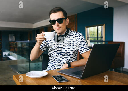 Junge hübsche Hipster Mann sitzt im Café Tasse Kaffee halten. Laptop und Handy auf Holztisch. Stockfoto