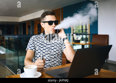 Junge hübsche Hipster Mann mit Bart sitzen im Café bei einer Tasse Kaffee, dampfen und Veröffentlichungen eine Wolke aus Vaporsitting. Am Laptop arbeiten und mit Stockfoto