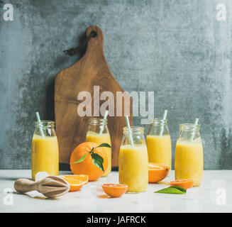 Gesunde gelbe Smoothie mit Zitrusfrüchten, Ingwer, Eis in Flaschen auf leichte Marmortisch, Holzbrett und graue Wand im Hintergrund, Tiefenschärfe, co Stockfoto