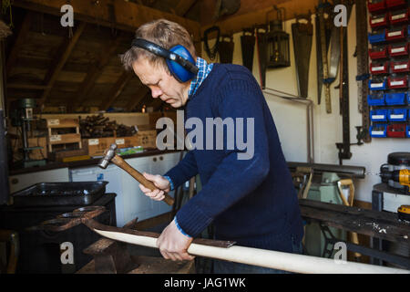 Mann, der in einer Werkstatt, tragen von Gehörschutz, mit einem Hammer auf ein Stück Holz arbeiten. Stockfoto