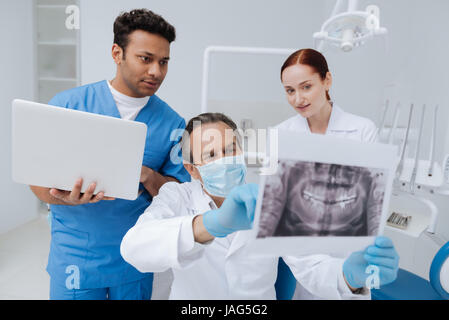 Schauen Sie aufmerksam. Kompetenter Zahnarzt trägt Maske auf seinem Gesicht und hält Bild des Kiefers beim sitzen zwischen zwei Assistenten Stockfoto
