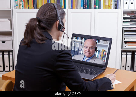 Frau mit Kopfhörer vor ihrem Laptop etwas auf ein Papier zu schreiben, dabei ein live video-Gespräch mit einem Kunden oder Kollegen kopieren Raum Stockfoto