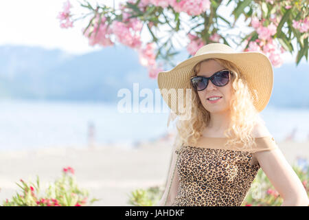 Schöne Blondine am Strand mit Hut und eine Sonnenbrille im Urlaub zwischen Oleander Blumen Stockfoto