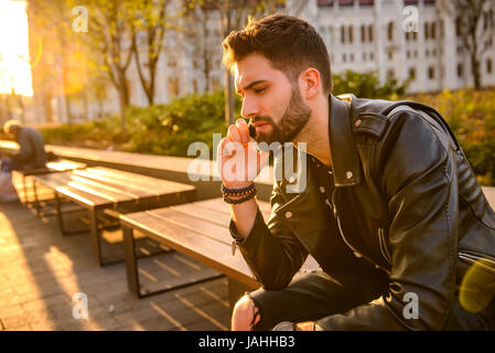 Porträt eines Mannes hübscher Junge Hipster in den Sonnenuntergang auf einer Bank sitzen und reden auf seinem Handy hautnah Stockfoto