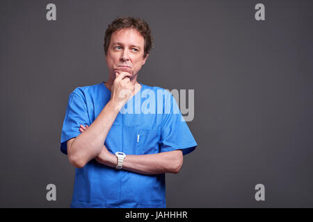 Einen mittleren Alters männlichen Arzt stehen und denken in einer blauen scheuert. Stockfoto