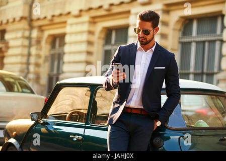Ein hübscher junger Geschäftsmann neben seinem Auto steht und mit seinem Handy Stockfoto