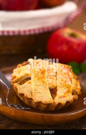 Kleine Runde Apfelkuchen mit Gitter Kruste auf Holzplatte mit Kuchengabel, in den Rücken, einen Apfel und ein Korb mit Äpfeln (selektiven Fokus, Fokus, ein Drittel in den Pie) Stockfoto