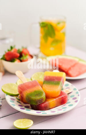 einige verschiedene hausgemachte natürliche Eis am Stiel in einer keramischen Platte neben einige verschiedene Stücke von Obst, wie Erdbeeren oder Wassermelone, auf einem rustikalen pi Stockfoto