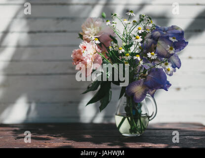 Bouquet von weißen Pfingstrosen, Kamillen und Iris Blumen in Glasvase. Sommer Hintergrund. Getönte Foto Stockfoto