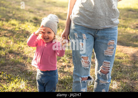 Spielendes Kind mit Mutter Wandern zusammen mit halten Hände im Sommerpark auf Rasen. Hauptthema ist Kind. Nicht erkennbare Mutter auf Foto.  Konzept Stockfoto