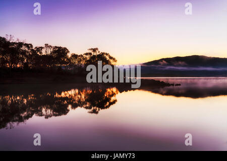 Dunklen kalten nebligen Sonnenaufgang am Lake Jindabyne im Winter. Der größte See in verschneiten Bergen reflektiert umliegenden Hügelketten und Gumtrees. Stockfoto