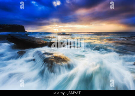 Stürmischen farbenfrohen Sonnenaufgang am Strand von Sydney Nordstrände Bungan. Rasche Waver fließt über Sandstein Felsen. Stockfoto