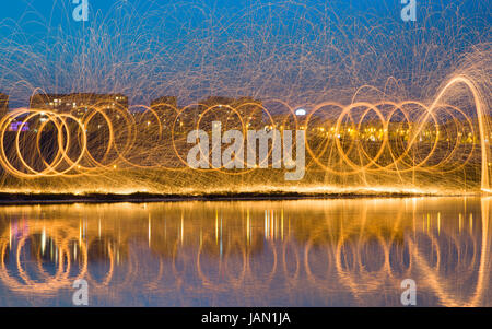 Heißen goldenen Funken fliegen vom Mann Spinning brennen Stahlwolle Fluss mit Wasserreflexion. Lange Belichtung Fotografie mit Stahlwolle brennen Stockfoto