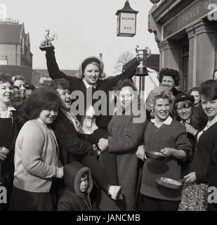 1965, historische, Bild zeigt die preisgekrönte Lady ihre Trophäen außerhalb der Prince Of Wales Pub in der jährlichen Pfannkuchen-Rennen auf der Old Kent Road, London, SE1 hochhalten. Stockfoto