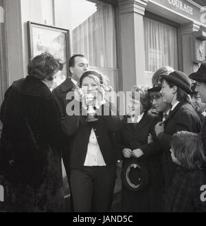 1965, zeigt historische, eine junge Frau ihre Trophäe, die aufgeregt lokale Schulkinder nach dem Gewinn der jährlichen Pfannkuchen Rennen auf der Old Kent Road, Peckham, London, zwischen dem Lord Wellington und Prince Of Wales Pubs. Stockfoto