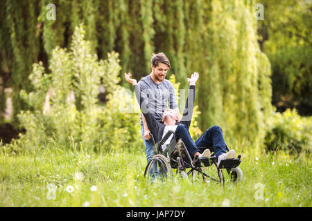 Hipster-Sohn mit behinderten Vater im Rollstuhl im Park spazieren. Stockfoto