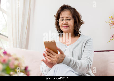 Glücklich senior Asiatin auf Sofa sitzen und mit einem smart-phone Stockfoto