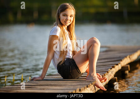 Lächelnde Mädchen sitzen am Pier in der Nähe von Teich im Sonnenuntergang Balken Stockfoto