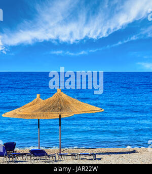 Sommer morgen kiesiger Strand mit Sonnenliegen und Sonnenschirmen strohig (Borsh, Albanien). Blauer Himmel mit Wolken. Stockfoto