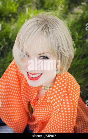 Close-up Portrait im freien junge schöne Mädchen in einer orange Hipster blonde helle fröhliche Tupfen Bluse, lächelnd rote volle Lippen auf die Zeitmessung Stockfoto