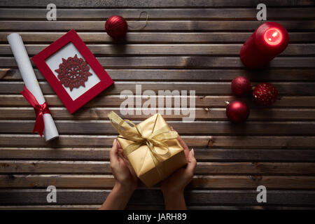 Bild von roten Schneeflocke in Frame, dekorative Spielzeug Kugeln, brennende Kerze und gewalzten Papier umwickelt mit rotem Seidenband rund um Geschenkbox in weiblichen Händen auf hölzernen Hintergrund Stockfoto