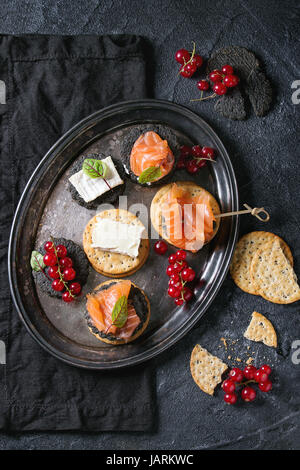 Stapel von schwarzen Holzkohle und traditionellen Cracker mit Räucherlachs, Frischkäse, grüner Salat und rote Johannisbeere Beeren auf Vintage Metalltablett über blac Stockfoto