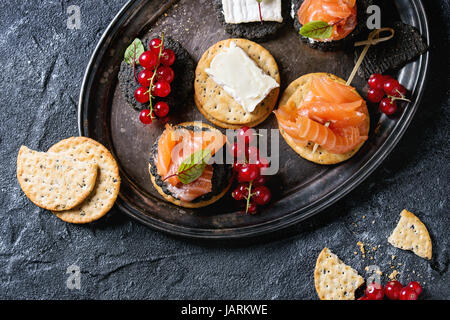 Stapel von schwarzen Holzkohle und traditionellen Cracker mit Räucherlachs, Frischkäse, grüner Salat und rote Johannisbeere Beeren auf Vintage Metalltablett über blac Stockfoto