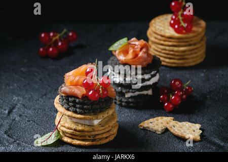 Stapel von schwarzen Vollkorn Holzkohle und traditionellen Cracker mit Räucherlachs, Frischkäse, grüner Salat und rote Johannisbeere Beeren über schwarzen Stein bac Stockfoto