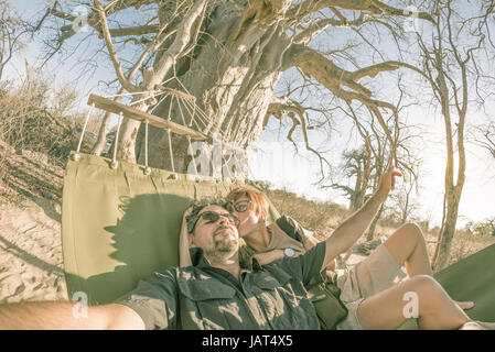 Paar nehmen Selfie auf Hängematte hängen von riesigen Baobab-Baum in der afrikanischen Savanne. Fisheye Ansicht, getönten Bild Hintergrundbeleuchtung. Wildnis Safari und adv Stockfoto