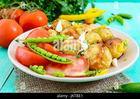 Würziger Salat aus gebackenen Kartoffeln, junger Knoblauch, Tomaten, grüne Erbsen auf einem Teller auf einem hellen Hintergrund. Stockfoto