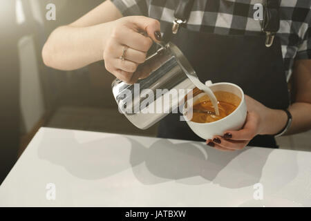 Barista strömenden Creme aus einem Krug in eine Tasse Kaffee, hielt sie in seinen Händen, stehend in der Sonne, Nahaufnahme Foto. Horizontalen Rahmen Stockfoto