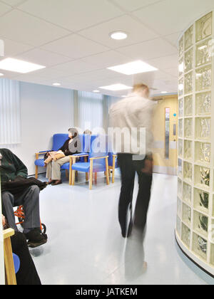 Patienten in NHS Krankenhaus Wartebereich, sitzend, wie ein Arzt durch den Rahmen, verschwommen durch Bewegung geht