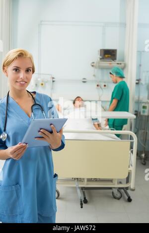 Blonde Krankenschwester hält einen Tablet-Computer neben einem Bett Stockfoto