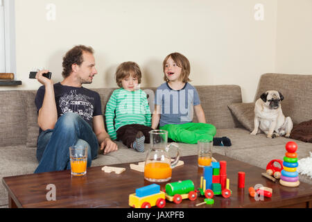 Vater mit Söhnen und Hund auf dem Sofa, im Vordergrund auf einem Tisch Spielzeug und Getränke Stockfoto