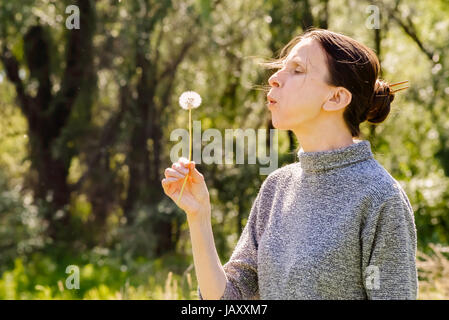 Erwachsene Frau bläst die Samen einer Löwenzahn Blume am Ende des Frühlings in der Nähe des Waldes Stockfoto