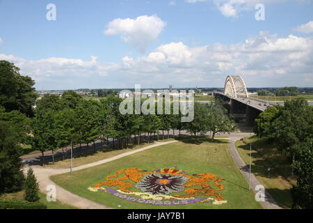 Blick über den Fluss Waal mit Waalbrug Bogenbrücke (1936), Nijmegen, Gelderland, Niederlande. Gesehen von Valkhof / Belvédère. Wappen der Stadt in den Blumen. Stockfoto