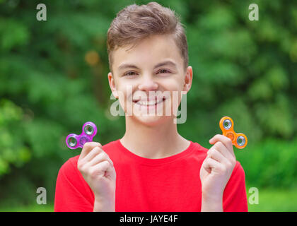 Junge teen boy Holding beliebte zappeln Spinner toy - draußen Portrait. Glücklich lächelnde Kind spielt mit einer orange Spinner im Sommer Park. Stockfoto