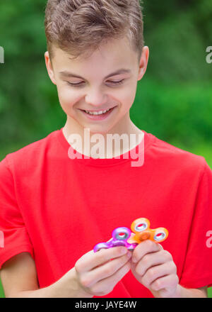 Junge teen boy Holding beliebte zappeln Spinner toy - draußen Portrait. Glücklich lächelnde Kind spielt mit einer orange Spinner im Sommer Park. Stockfoto