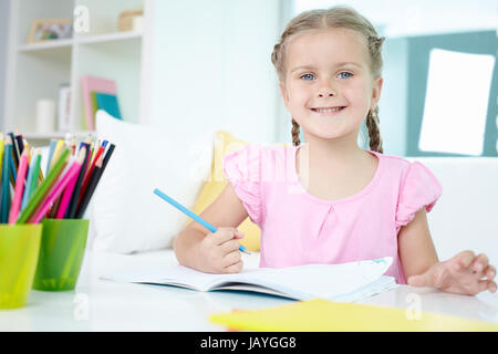 Niedliche kleine Mädchen vom Tisch mit Buntstiften und offene Copybook vor ihr sitzt Stockfoto